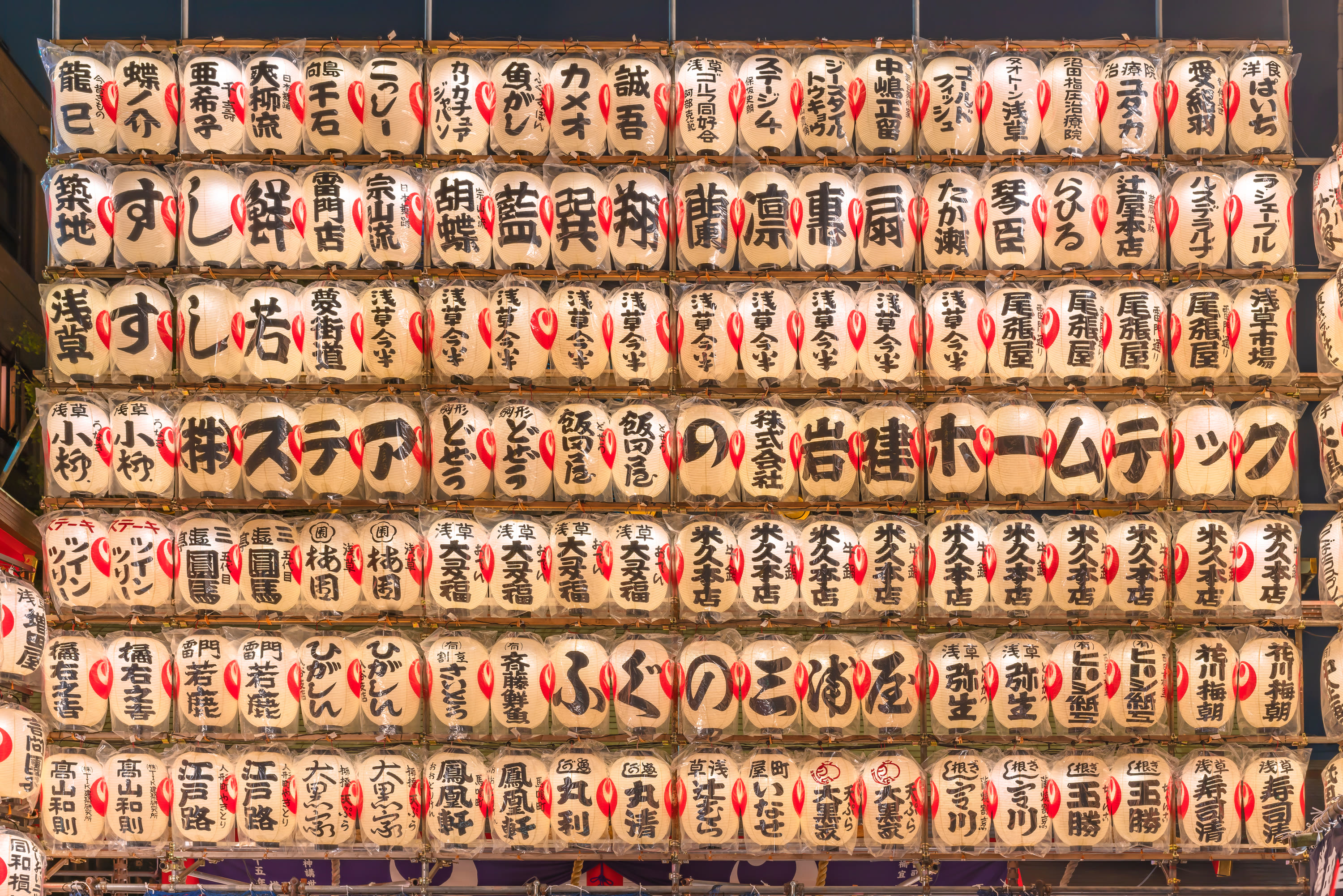 A wall of glowing paper lanterns with sponsors' names brushed in black ink