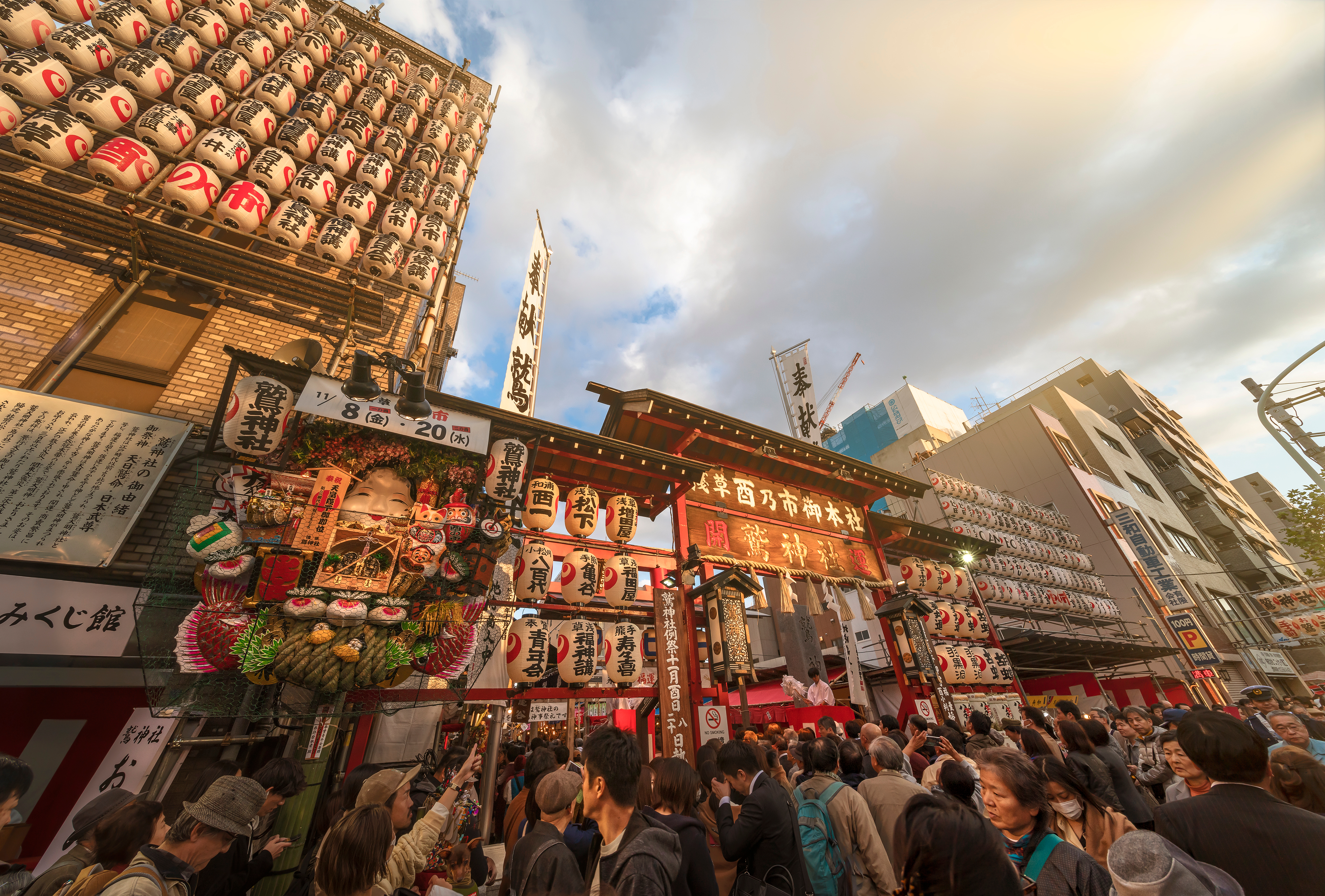 Daytime crowd at the Otori Shrine main gate with a large decorated rake, lanterns and the Tori no Ichi signboard