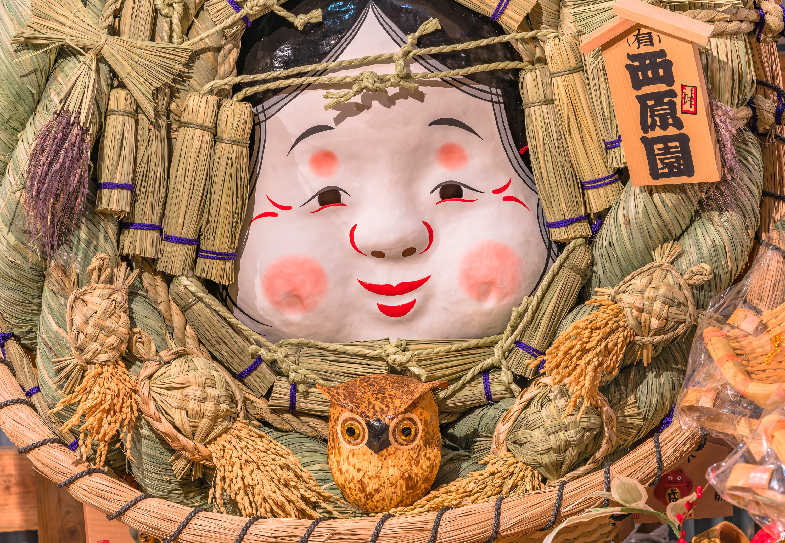 Smiling Okame face at the center of a giant straw kumade, with an owl ornament below