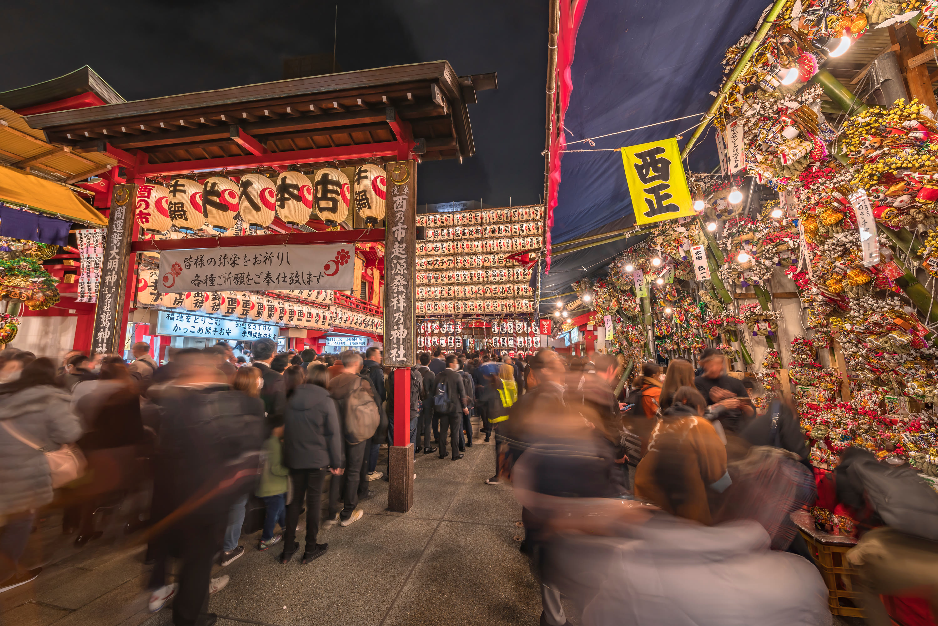 Night crowd lining up under the Otori Shrine gate with paper lanterns, kumade rake stalls packed on the right